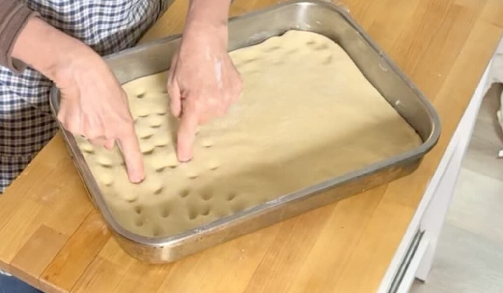 A person presses their fingers into rectangular German butterkuchen dough in a baking tray, creating dimples on the surface. The tray rests on a wooden countertop.