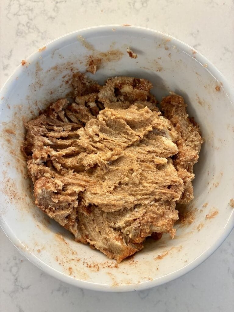 A white bowl filled with cinnamon-swirled dough sits on a light marble countertop. The dough appears soft and partly mixed, with visible streaks of cinnamon throughout.
