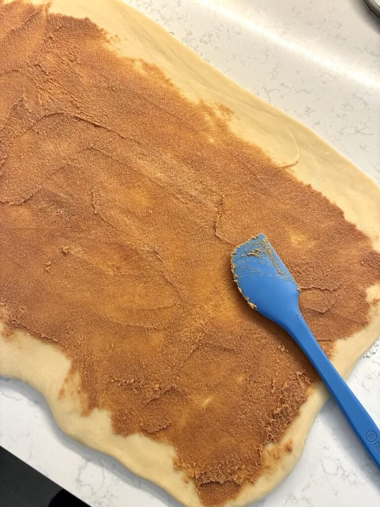 A rolled-out dough is spread with a cinnamon-sugar mixture. A blue spatula rests on the dough, showing the spreading process on a white countertop.