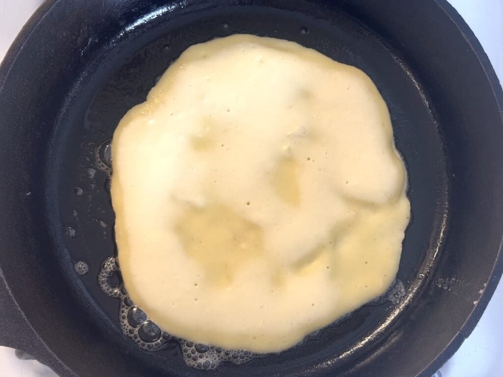 A sourdough pancake cooking in a black skillet, with bubbles forming on the surface and edges beginning to set.