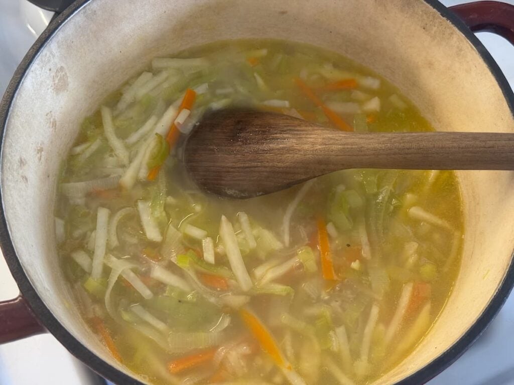 A pot of vegetable soup with julienned carrots, celery, and potatoes simmering in broth, being stirred with a wooden spoon—perfect for pairing with traditional German pancake soup.