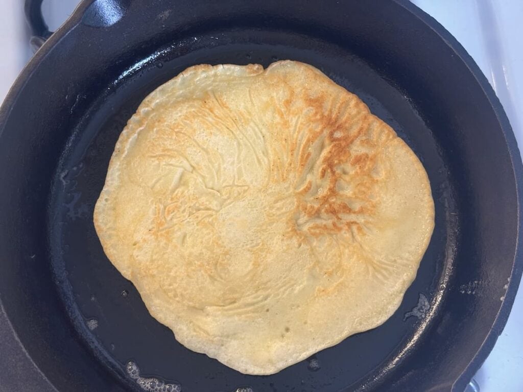 A golden-brown sourdough pancake cooking in a black cast iron skillet on a stovetop.