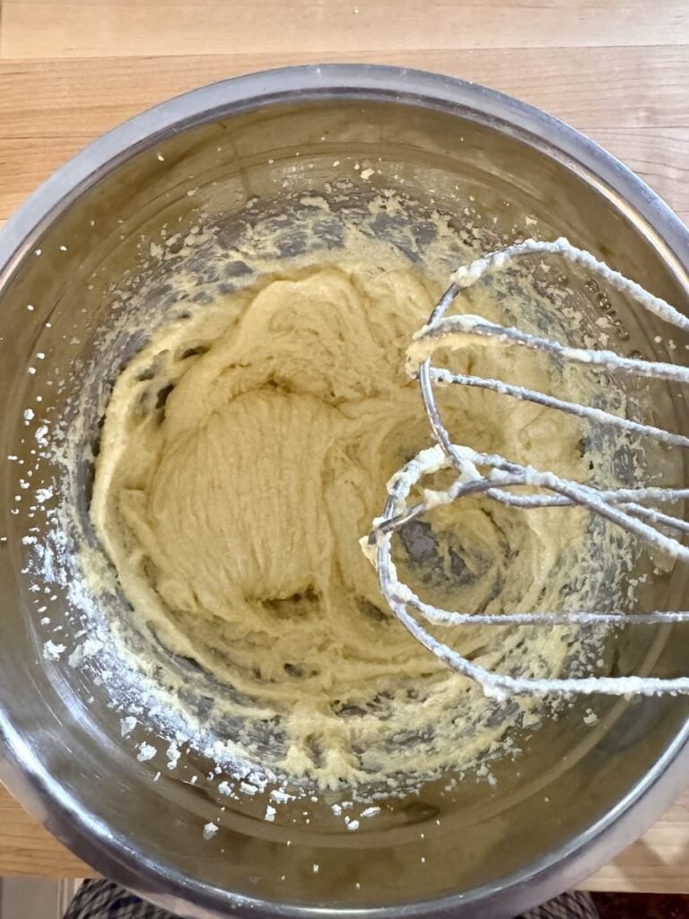A metal mixing bowl with creamy batter inside, shown from above. An electric mixer with two beaters, coated in batter, rests on the right side of the bowl. The bowl sits on a wooden surface.