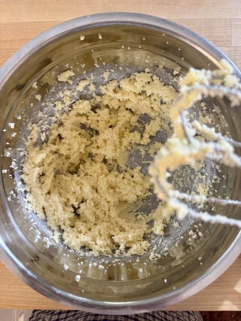 A metal mixing bowl with creamed butter and sugar inside, next to a hand mixer with some mixture on its beaters, placed on a wooden surface.