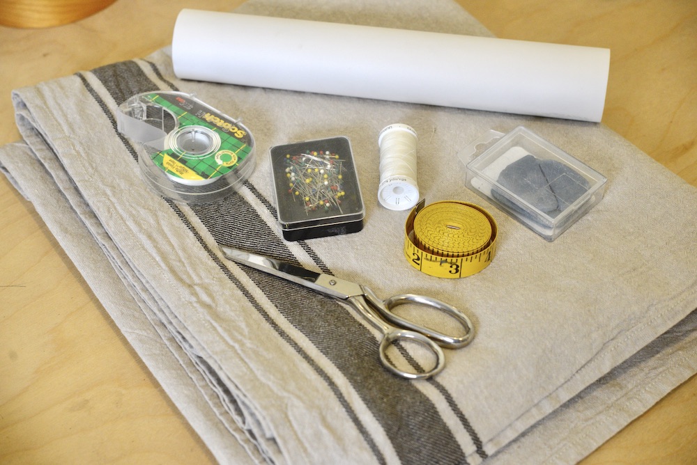 linen tablecloth, scissors, tailor's chalk, tape measure, thread, pins, scotch tape, and a roll of pattern paper on table