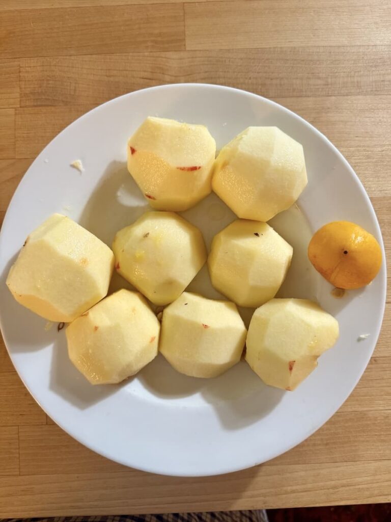 Eight peeled apples and one unpeeled apple are arranged on a white plate, which sits on a wooden surface.
