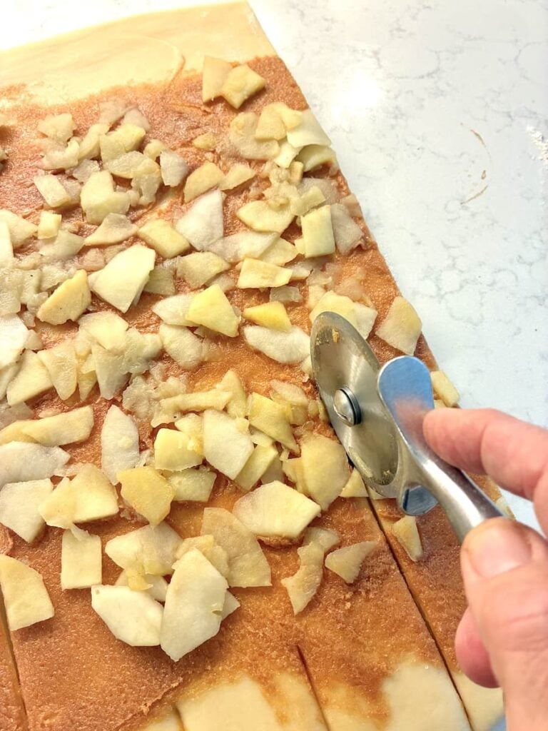 A hand uses a pizza cutter to slice dough topped with chopped apples and cinnamon sugar on a white countertop.