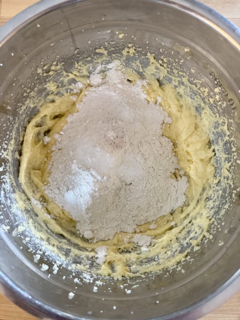 A metal mixing bowl containing yellow batter with a pile of white flour and sugar on top, being mixed together on a wooden surface.