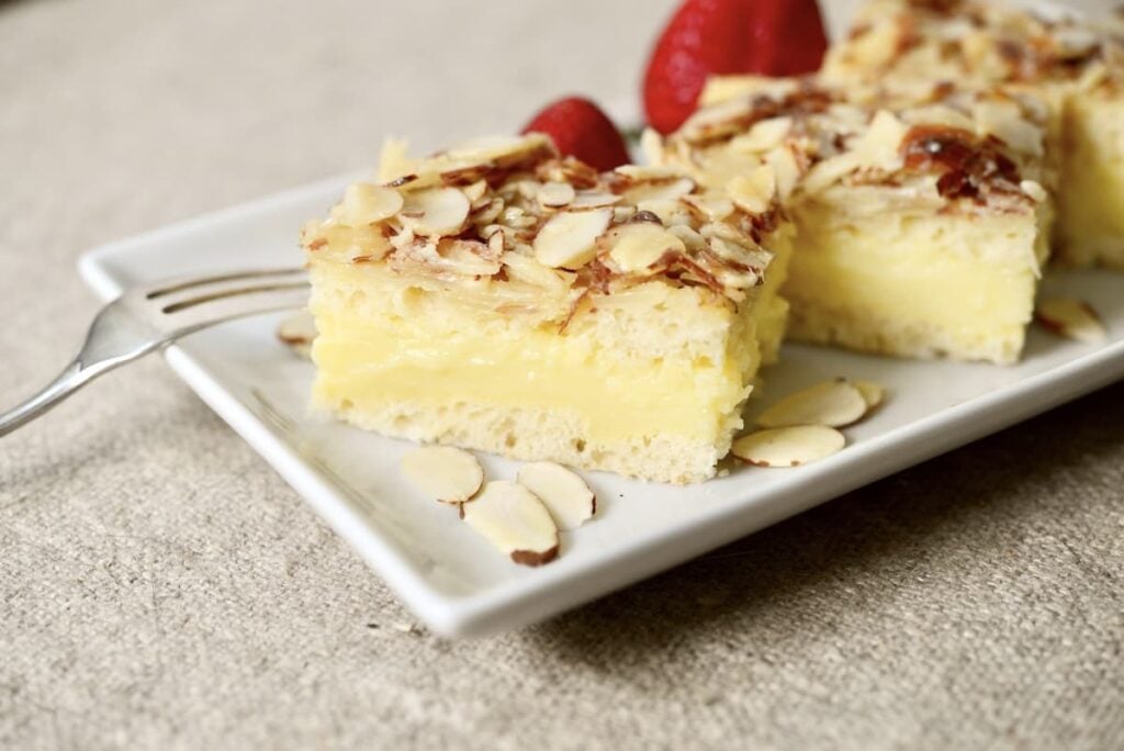 Two slices of custard-filled almond cake sit on a white plate, garnished with sliced almonds. A fork and two strawberries are also on the plate, all set on a beige fabric surface.
