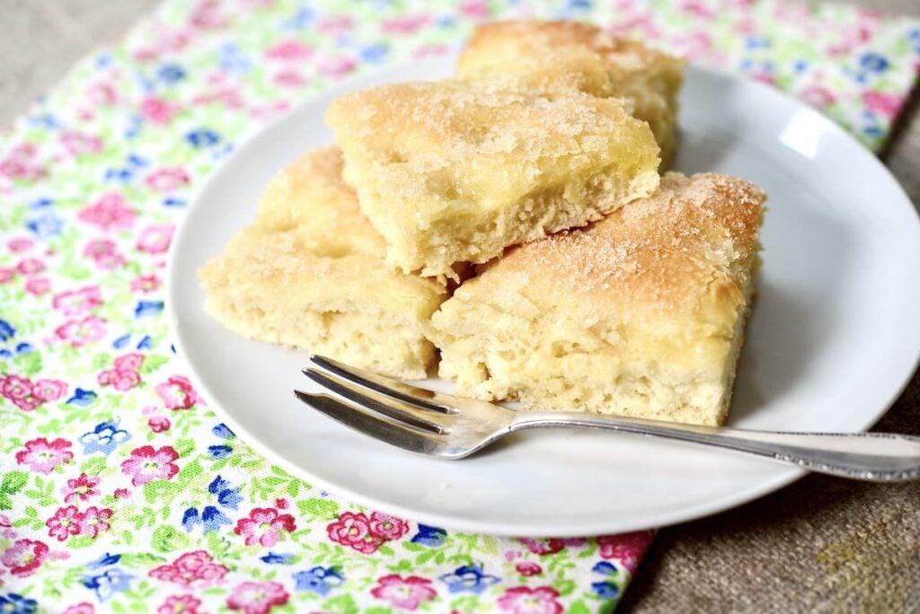 Four pieces of sugar-topped German butterkuchen are arranged on a white plate with a fork. The plate sits on a colorful, floral-patterned napkin atop a light surface.