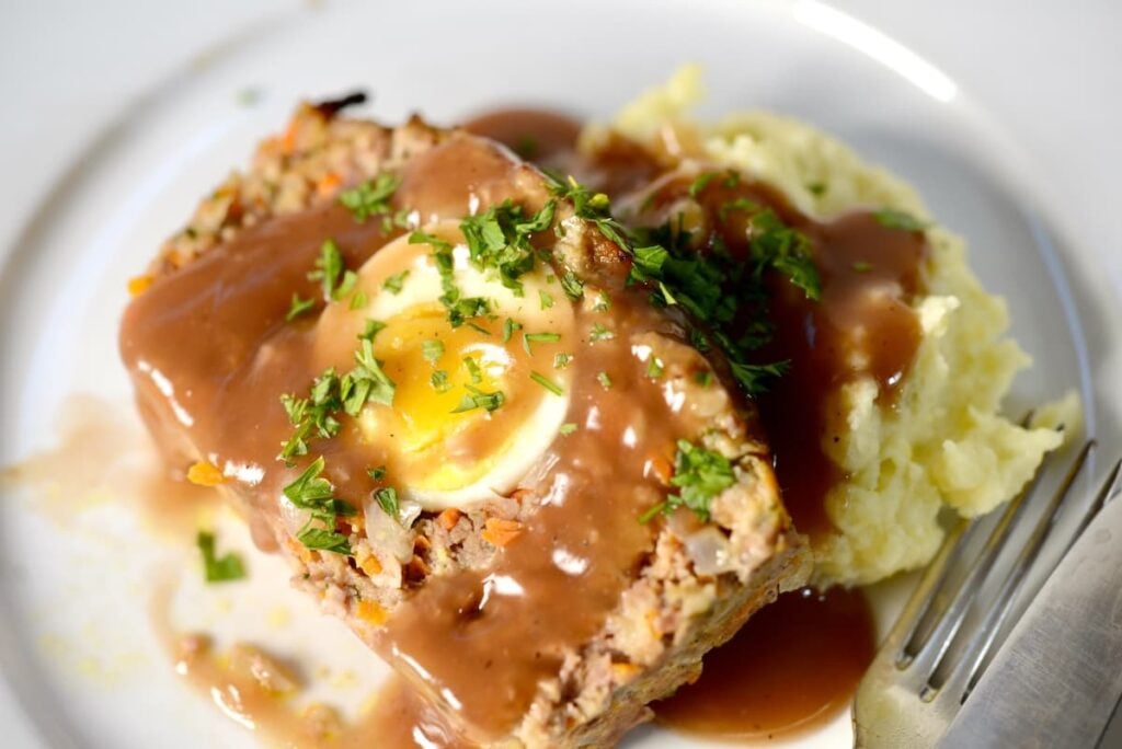 A slice of meatloaf containing a hard-boiled egg, topped with gravy and herbs, served with a side of mashed potatoes.