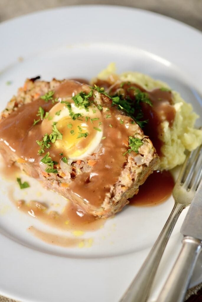 A slice of meatloaf containing a hard-boiled egg, topped with gravy and herbs, served with mashed potatoes on a white plate with a fork and knife.