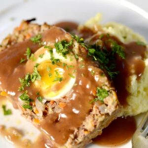 A slice of meatloaf containing a hard-boiled egg, topped with gravy and herbs, served with a side of mashed potatoes.