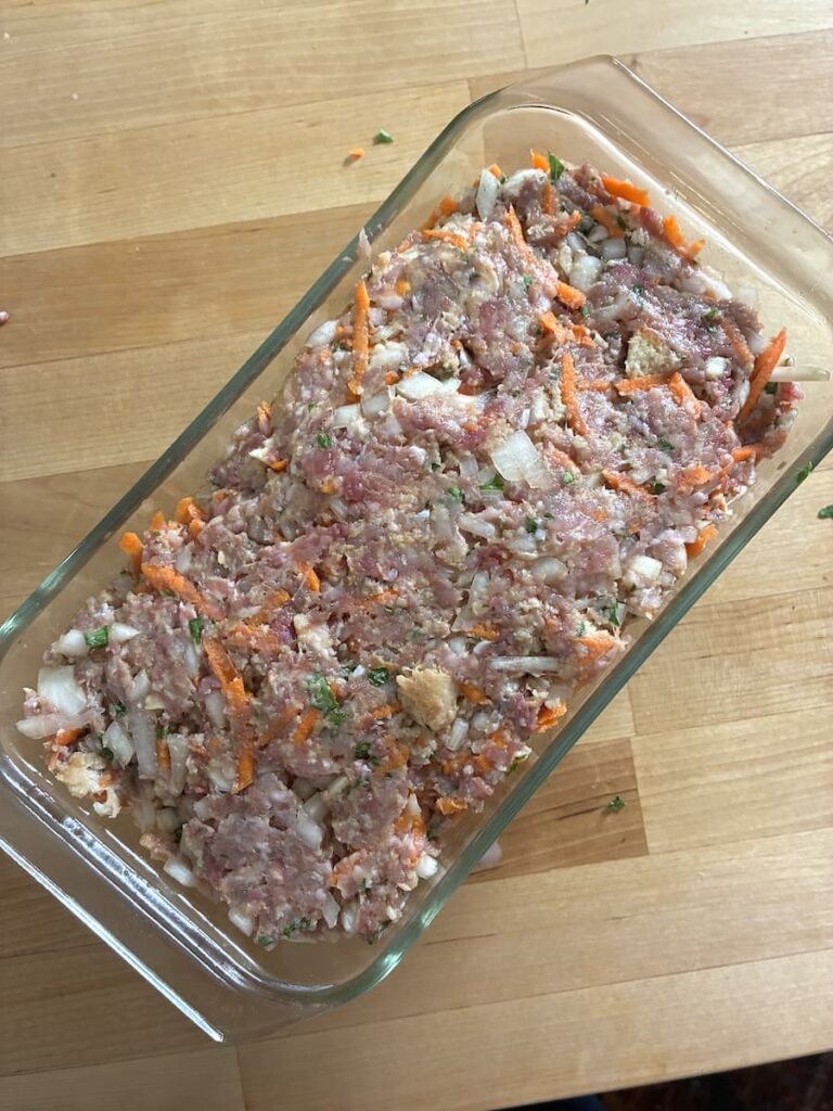 Raw meatloaf mixture with visible pieces of onion, carrot, and breadcrumbs in a glass loaf pan on a wooden surface.