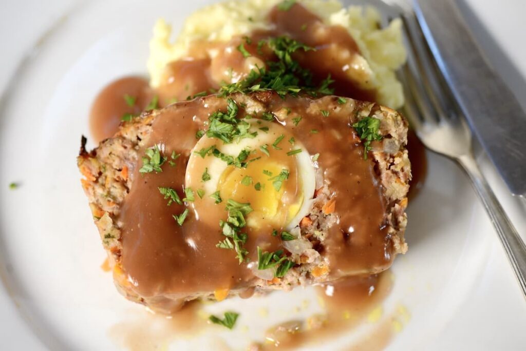 A slice of meatloaf with a hard-boiled egg in the center, covered in gravy and herbs, served with a side of mashed potatoes.
