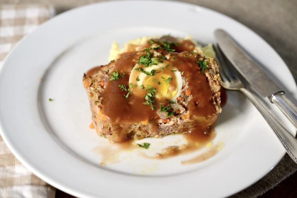 A slice of meatloaf containing a hard-boiled egg, topped with gravy and herbs, served on a white plate with a fork and knife.