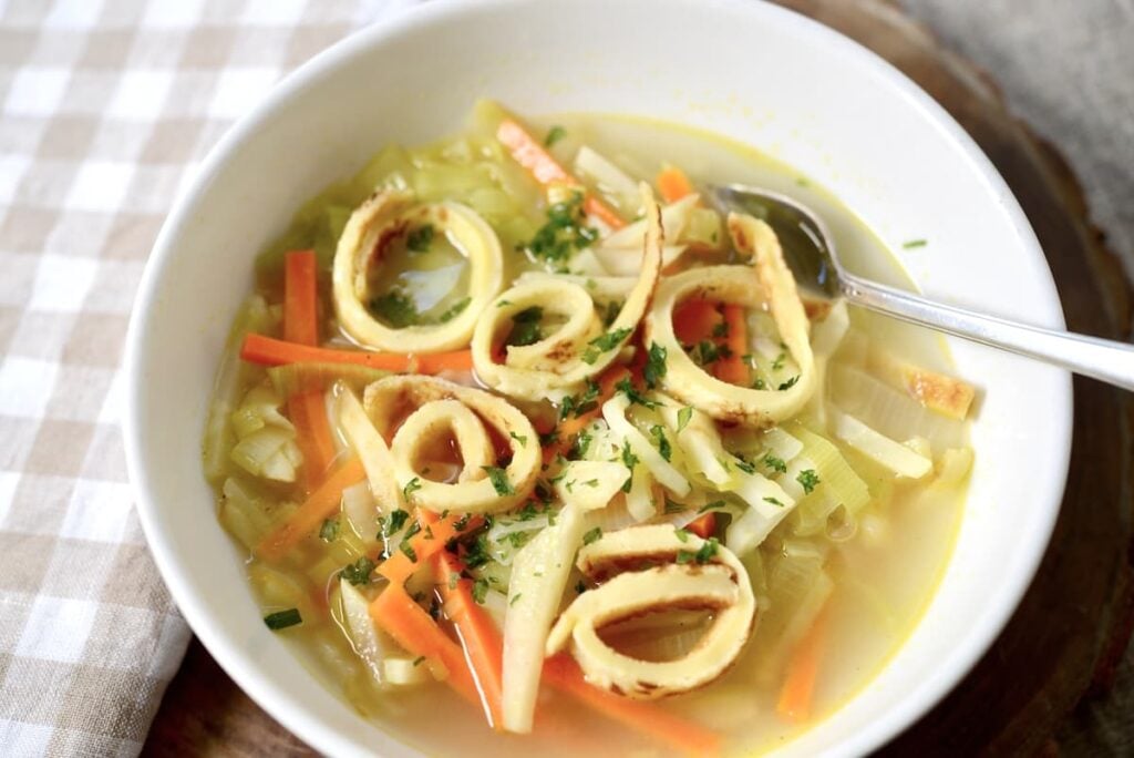 A bowl of clear German pancake soup with sliced vegetables and crispy fried rings, garnished with chopped herbs. A spoon rests inside the white bowl, which sits on a wooden surface next to a checkered cloth.