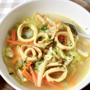 A bowl of clear German pancake soup with sliced vegetables and crispy fried rings, garnished with chopped herbs. A spoon rests inside the white bowl, which sits on a wooden surface next to a checkered cloth.