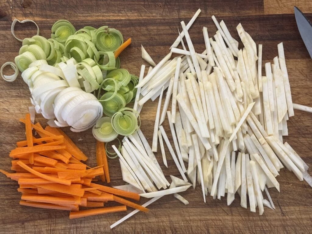 Sliced vegetables on a wooden cutting board: orange carrot sticks, round green and white leek slices, and thin strips of white root vegetable—ingredients often used in German pancake soup. A knife is partially visible on the right.
