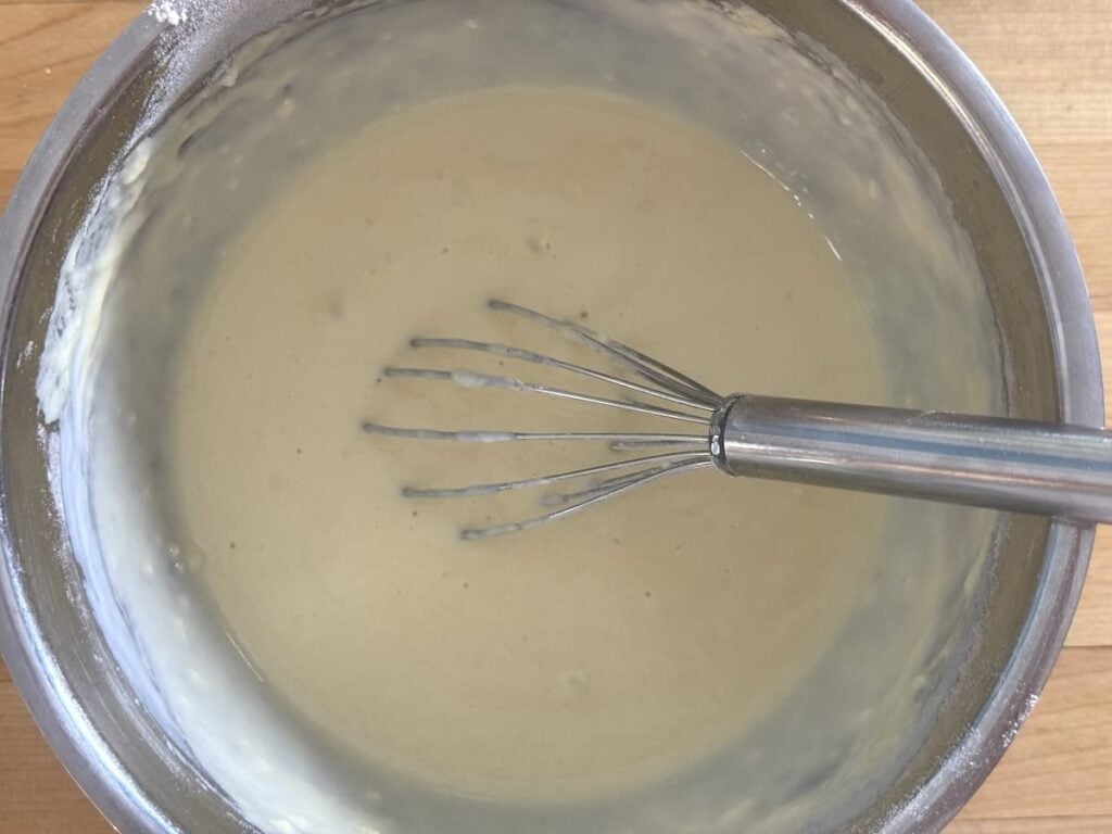 A metal whisk rests in a stainless steel bowl filled with smooth, light sourdough pancake batter, sitting on a wooden surface. Batter clings to the sides of the bowl, indicating it has just been mixed.