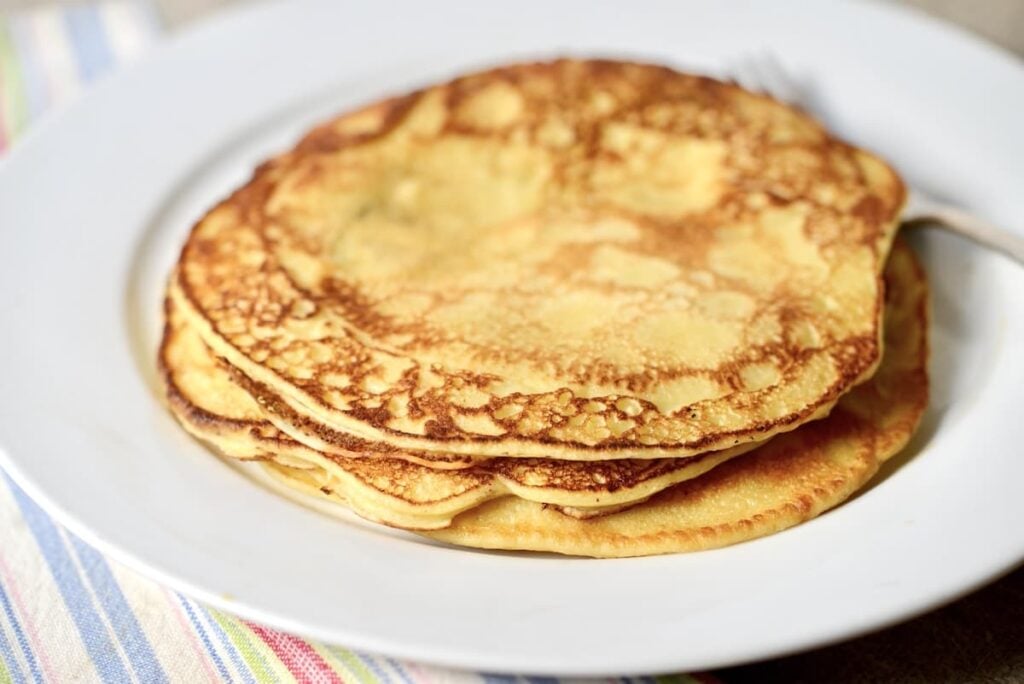 A stack of three golden-brown sourdough pancakes sits on a white plate with a fork beside them, placed on a striped cloth.