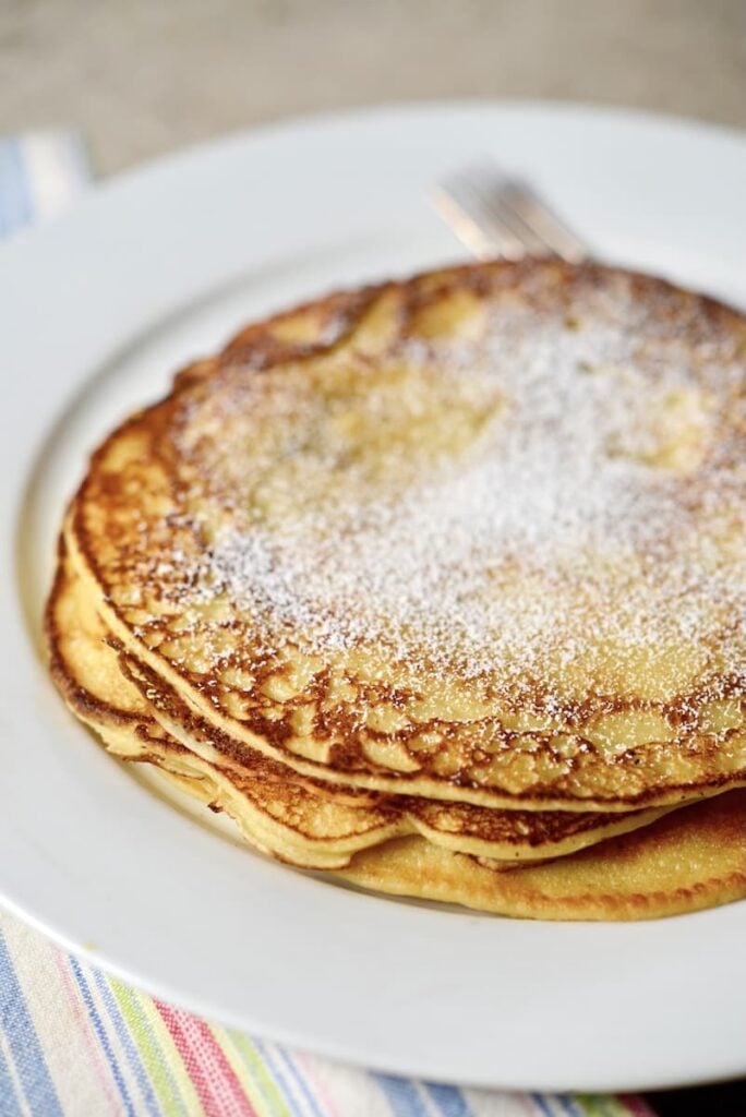 A stack of golden sourdough pancakes dusted with powdered sugar sits on a white plate. A fork rests on the plate, and a striped cloth is partially visible underneath.