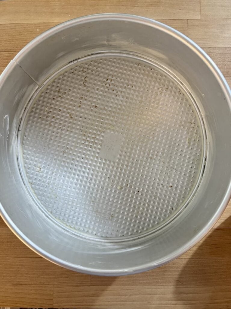 A greased round springform cake pan with a textured bottom, placed on a wooden surface.