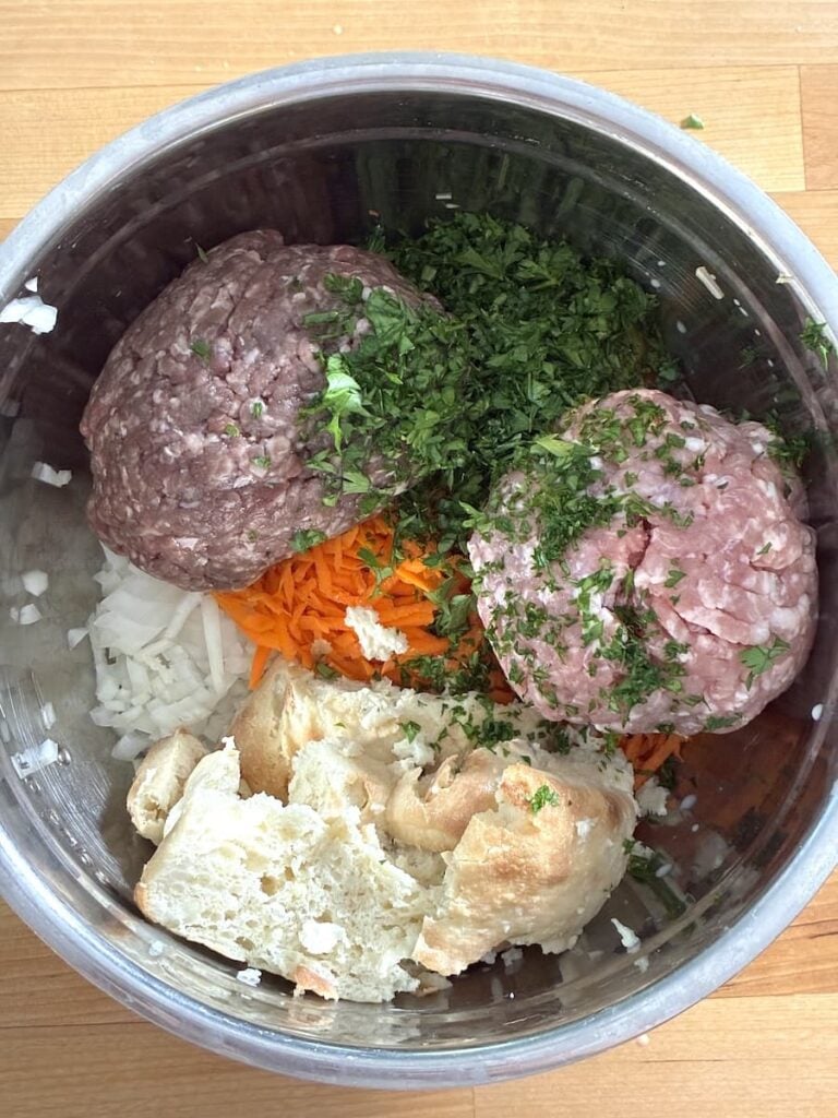A mixing bowl containing raw ground meat, chopped fresh herbs, shredded carrots, diced onions, and pieces of bread.