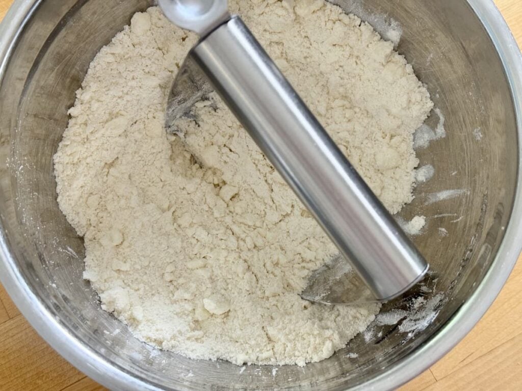 A metal pastry blender sits in a stainless steel bowl with flour and bits of butter, starting to mix the ingredients on a wooden surface.