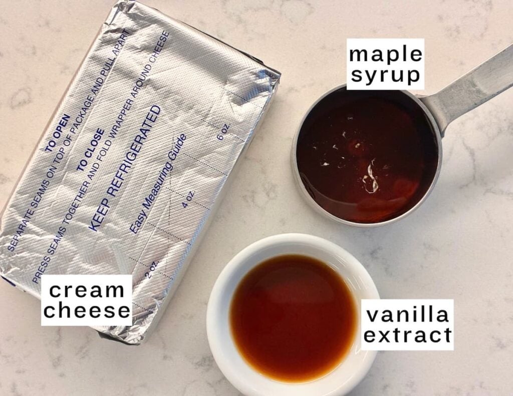A block of cream cheese, a measuring cup of maple syrup, and a small bowl of vanilla extract are arranged on a white countertop. Each ingredient is labeled with text.