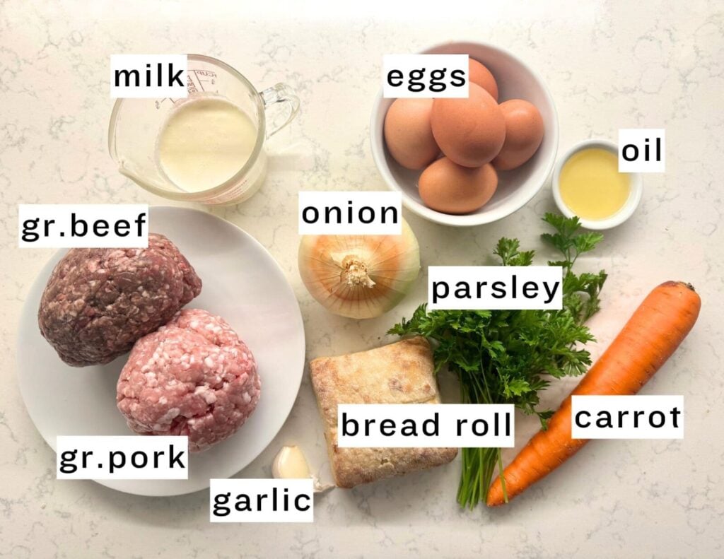 Ingredients for meatloaf: milk, ground beef, ground pork, garlic, onion, eggs, bread roll, parsley, oil, and carrot arranged on a countertop.