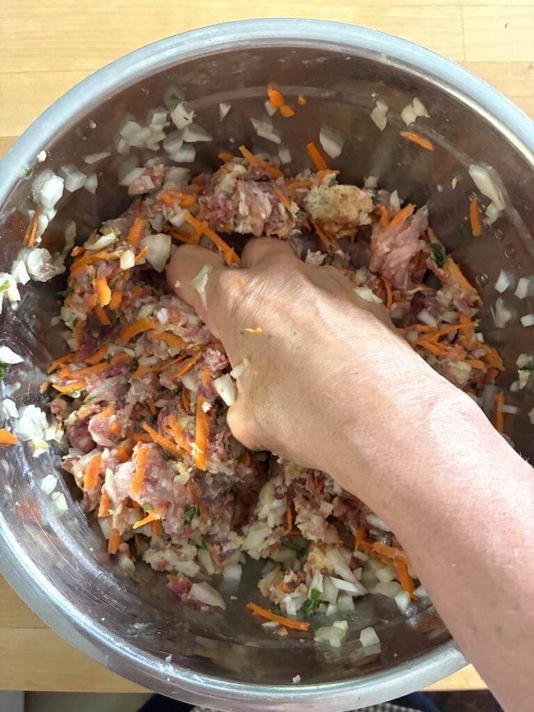 A hand mixes raw ground meat with chopped onions and shredded carrots in a stainless steel bowl.