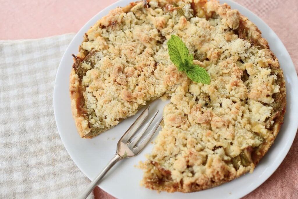 A crumb-topped pie with one slice missing sits on a white plate beside a fork. A sprig of mint garnishes the pie, and the plate rests on a checked cloth over a pink surface.