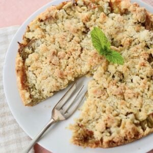 A crumb-topped pie with one slice missing sits on a white plate beside a fork. A sprig of mint garnishes the pie, and the plate rests on a checked cloth over a pink surface.