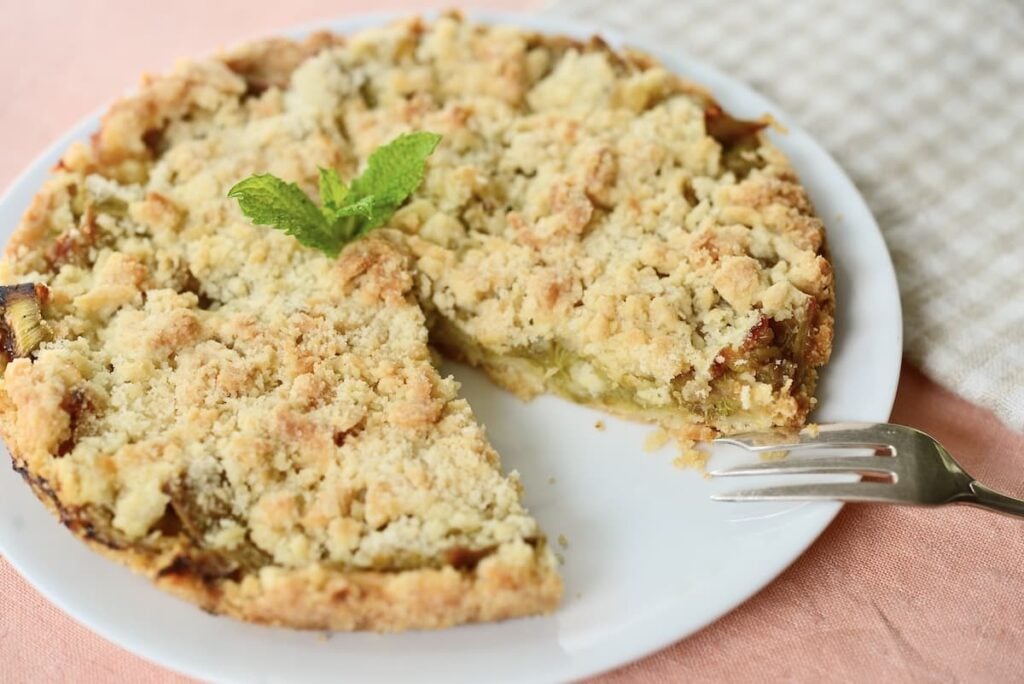 A crumbly pie with a missing slice sits on a white plate, garnished with a mint sprig. A fork rests beside the pie, and a checkered cloth is partially visible in the background.