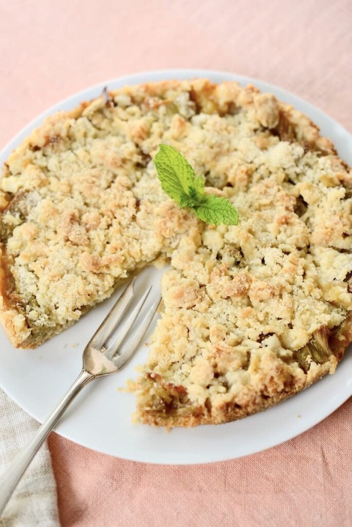 A round crumb-topped pie with a slice missing, garnished with a sprig of mint, sits on a white plate next to a fork on a light pink tablecloth.