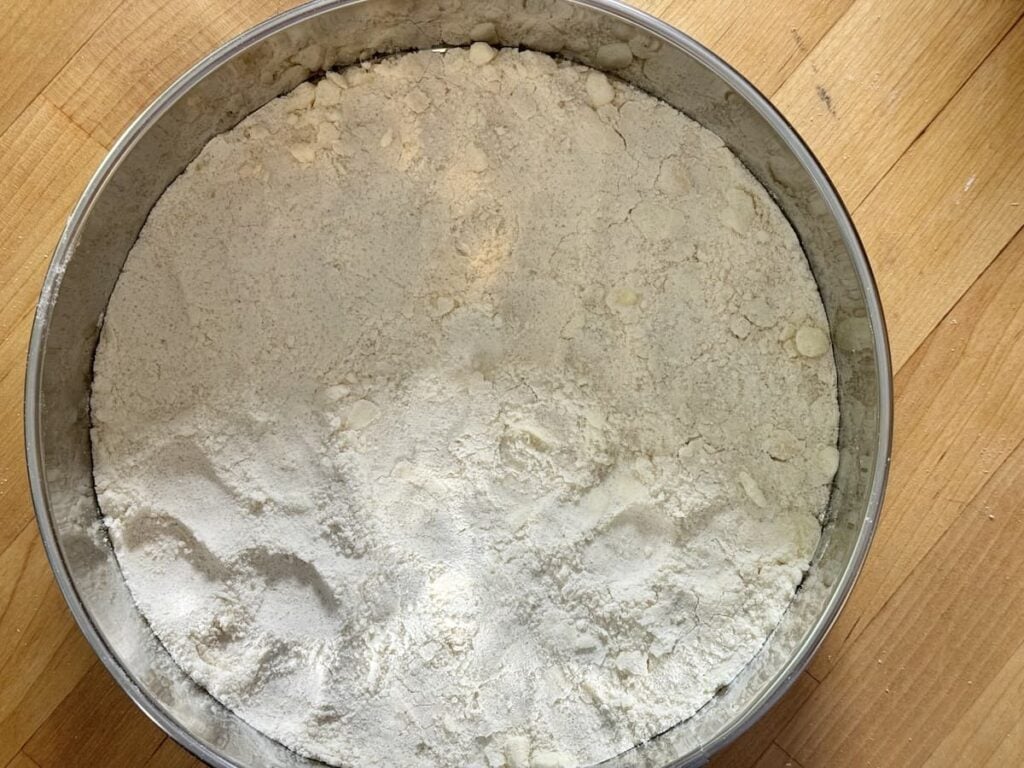 A round metal container filled with white flour sits on a light wooden surface. Some small lumps are visible in the flour.