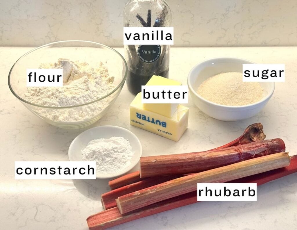 A display of labeled baking ingredients on a counter: a bowl of flour, a stick of butter, a bowl of sugar, cornstarch in a small dish, vanilla extract bottle, and several stalks of fresh rhubarb.
