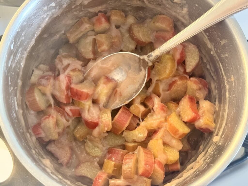 A close-up of a saucepan filled with cooked rhubarb pieces in a thick, pink sauce. A metal spoon rests in the pot, partially submerged in the mixture.