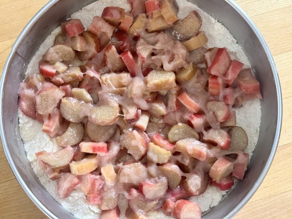 A round baking pan filled with sliced rhubarb on top of a layer of flour mixture, ready to be baked, placed on a wooden surface.
