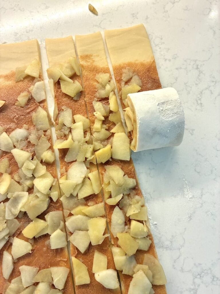 Four strips of dough topped with chopped apples are being prepared on a floured surface. One strip is partly rolled up into a spiral, ready to be baked, while the rest lie flat with apple pieces scattered on top.