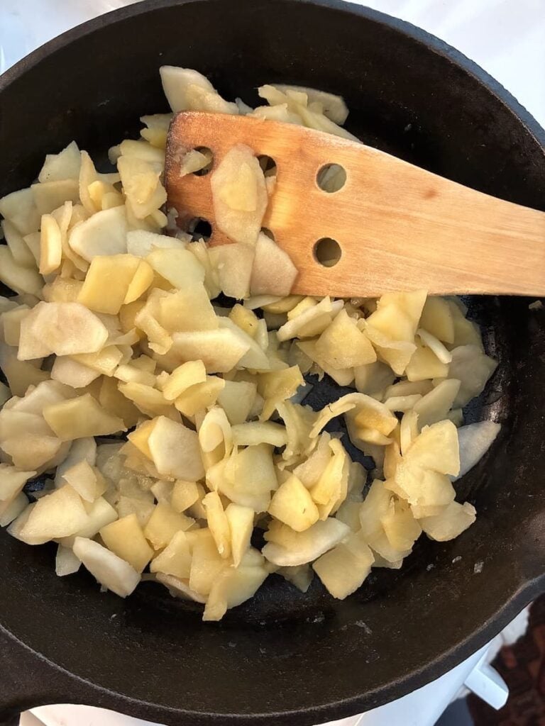 Chopped apples cooking in a black skillet, stirred with a wooden spoon.
