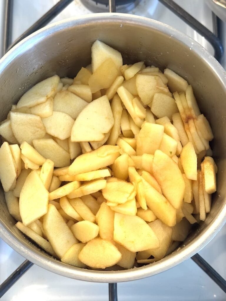 Thinly sliced apples are piled in a metal pot on a stove, ready to be cooked or baked for a delicious apple crumb cake.