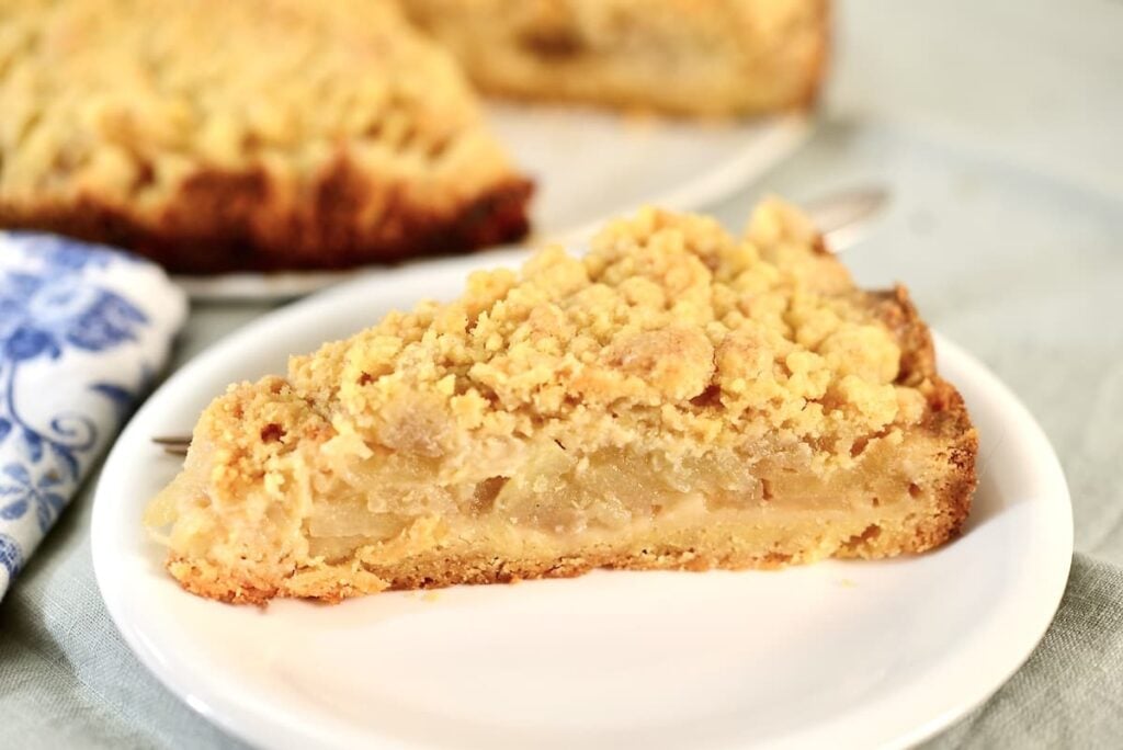 A slice of apple crumble pie with a golden, crumbly topping sits on a white plate, reminiscent of classic apple crumb cake, with more pie visible in the background.