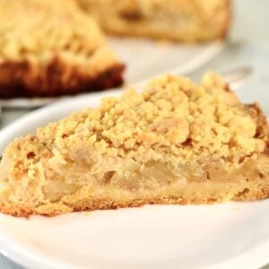 A slice of apple crumble pie with a golden, crumbly topping sits on a white plate, reminiscent of classic apple crumb cake, with more pie visible in the background.