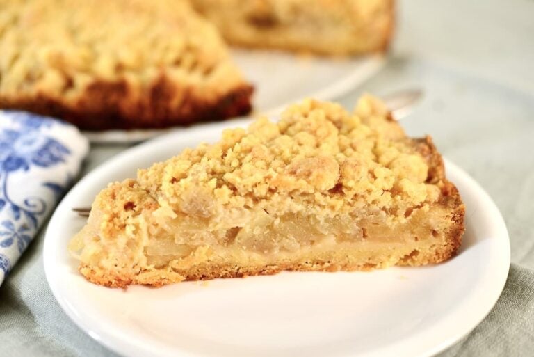 A slice of apple crumble pie with a golden, crumbly topping sits on a white plate, reminiscent of classic apple crumb cake, with more pie visible in the background.