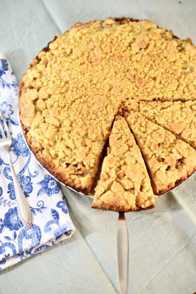 A round apple crumb cake pie with one slice being served on a pie server, next to a blue and white patterned napkin and two forks.