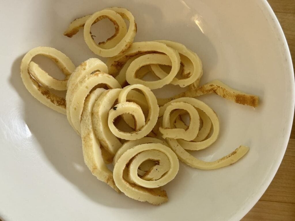 Several thin pancake spirals are arranged in a white bowl on a wooden surface, ready to be served as a traditional German pancake soup.