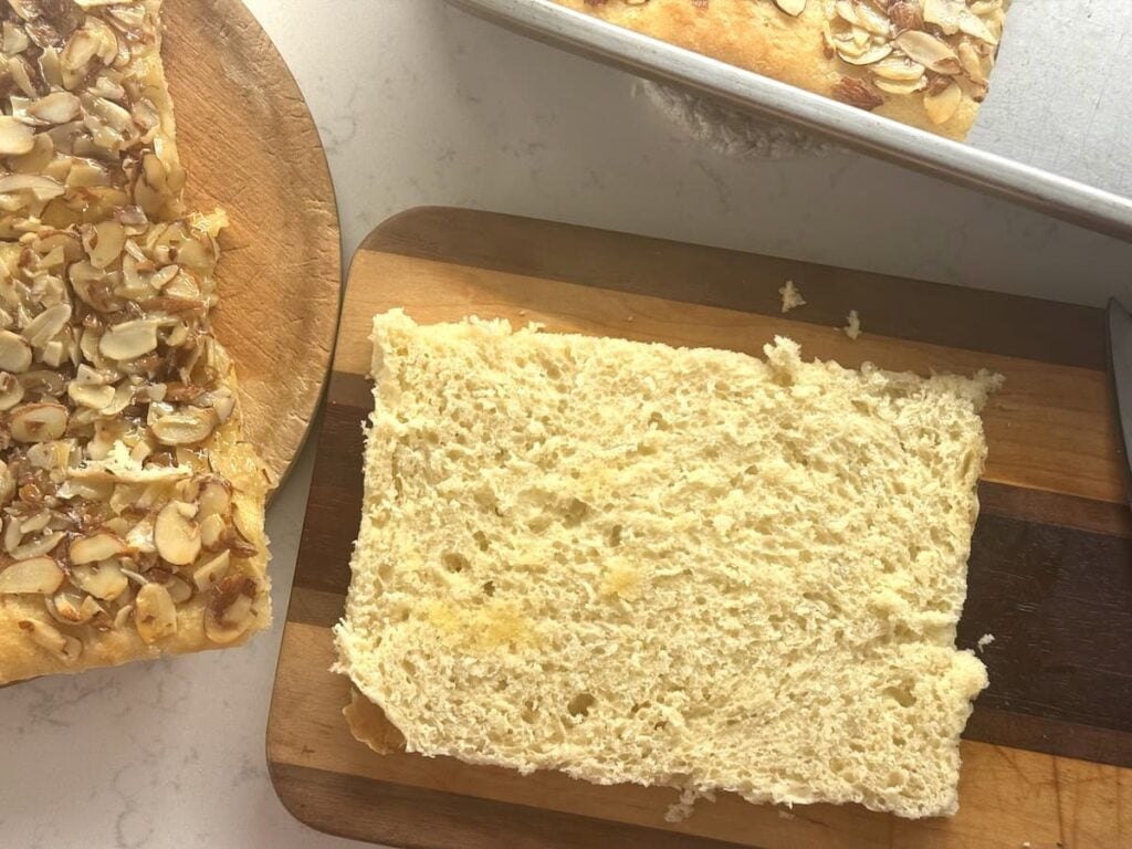 A thick slice of light, airy bread rests on a wooden cutting board, next to a larger piece of bread topped with slivered almonds, sitting on a round wooden board. A baking tray is partially visible in the corner.