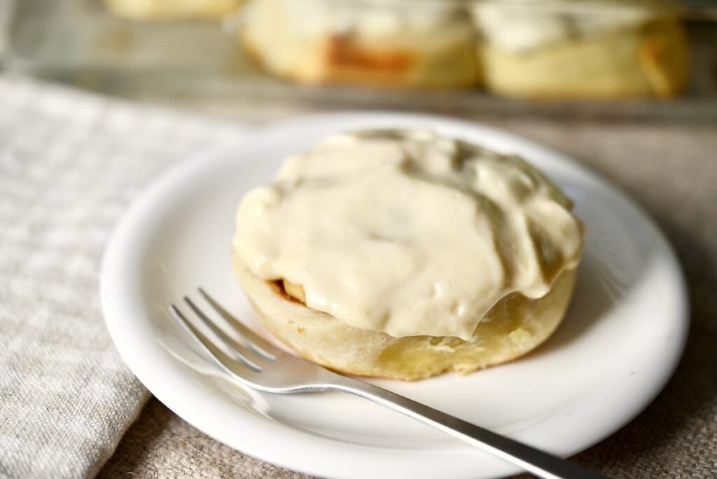 A cinnamon roll topped with thick white icing sits on a white plate next to a fork, with a cloth napkin and more cinnamon rolls blurred in the background.